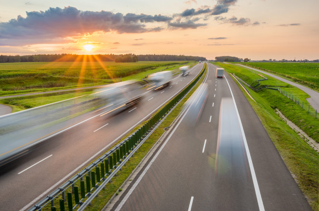 View of a highway with trucks and vehicles passing as the sun sets in the far horizon below clouds. 